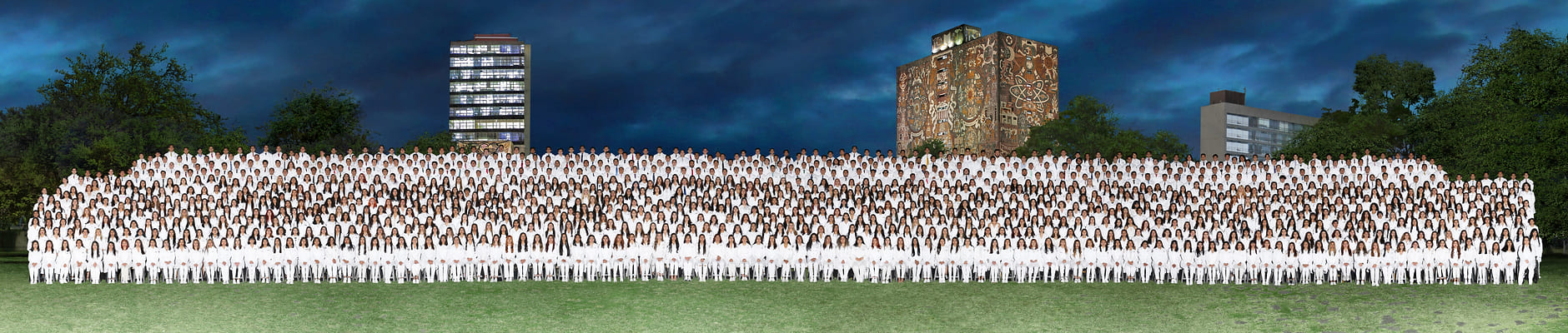 Foto panorámica de una generación completa de la UNAM, cientos de egresados formados en filas sobre la explanada de Ciudad Universitaria al anochecer, con la Torre de Rectoría al fondo.